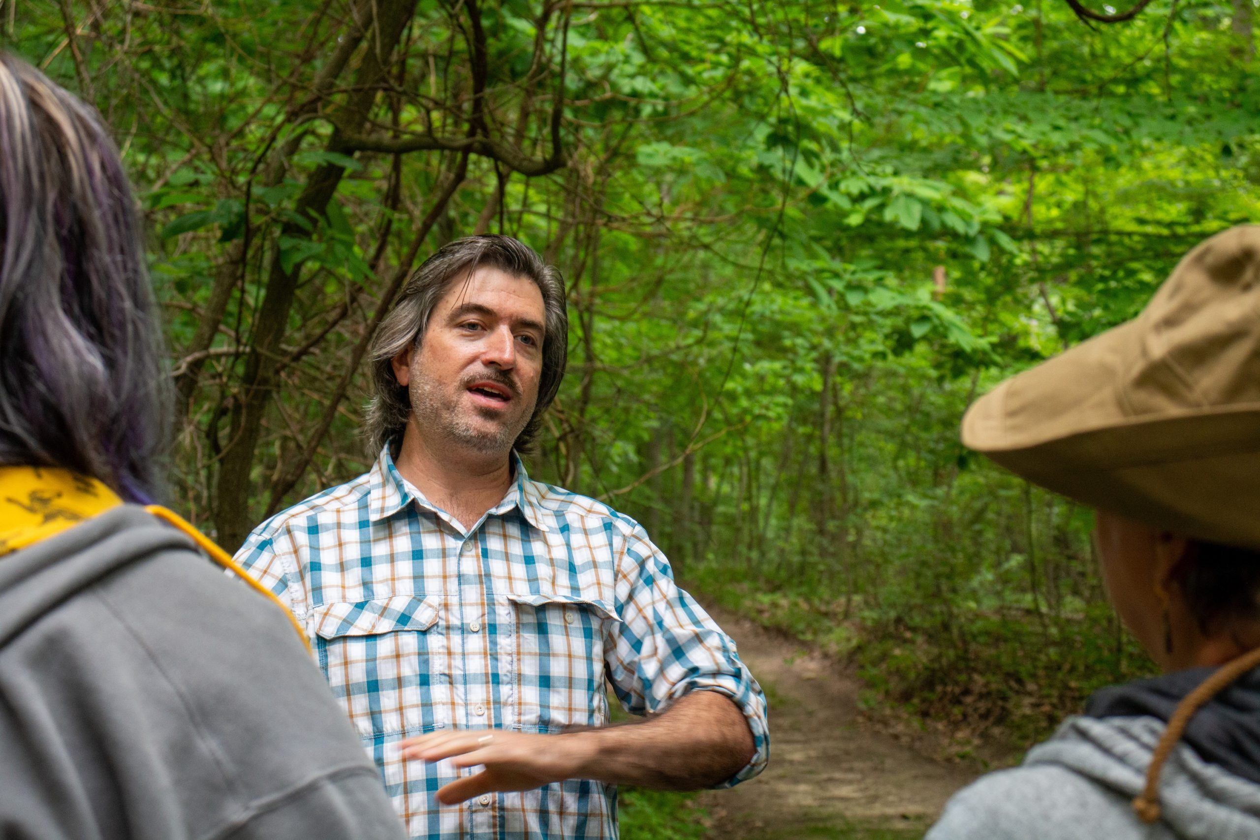 A man in a plaid shirt speaks to two student researchers from Alma College on a wooded trail during the Bog Event, surrounded by green trees and lush foliage. One listener wears a wide-brimmed hat; another has purple streaks in her hair.