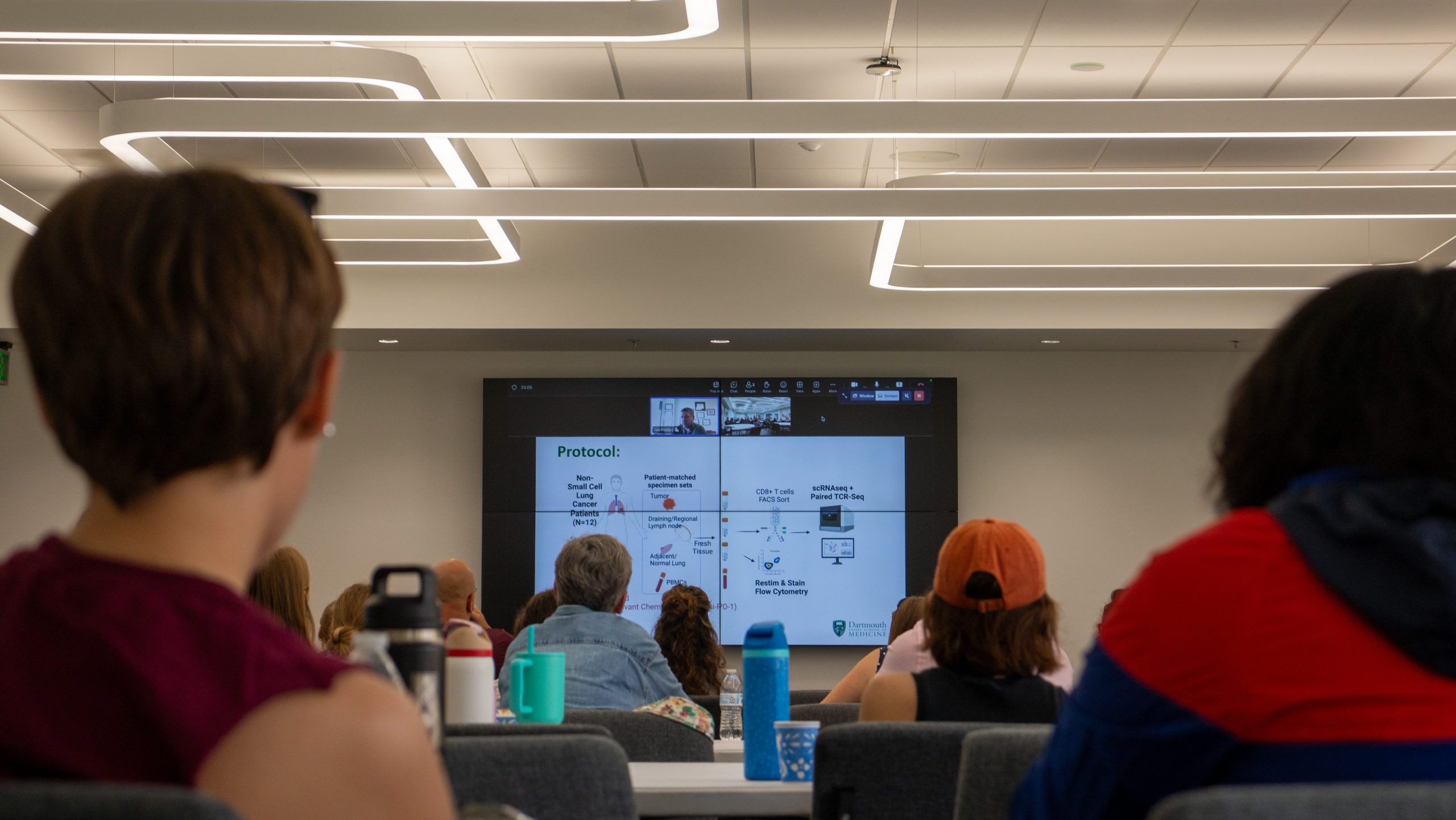 Students sit in a classroom facing a large screen displaying a presentation slide with diagrams and text. Modern ceiling lights illuminate the room, and various water bottles are on desks.