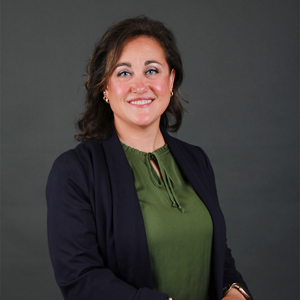 A woman with wavy brown hair wears a dark blazer over an olive green blouse. She is smiling and posed against a plain dark background.