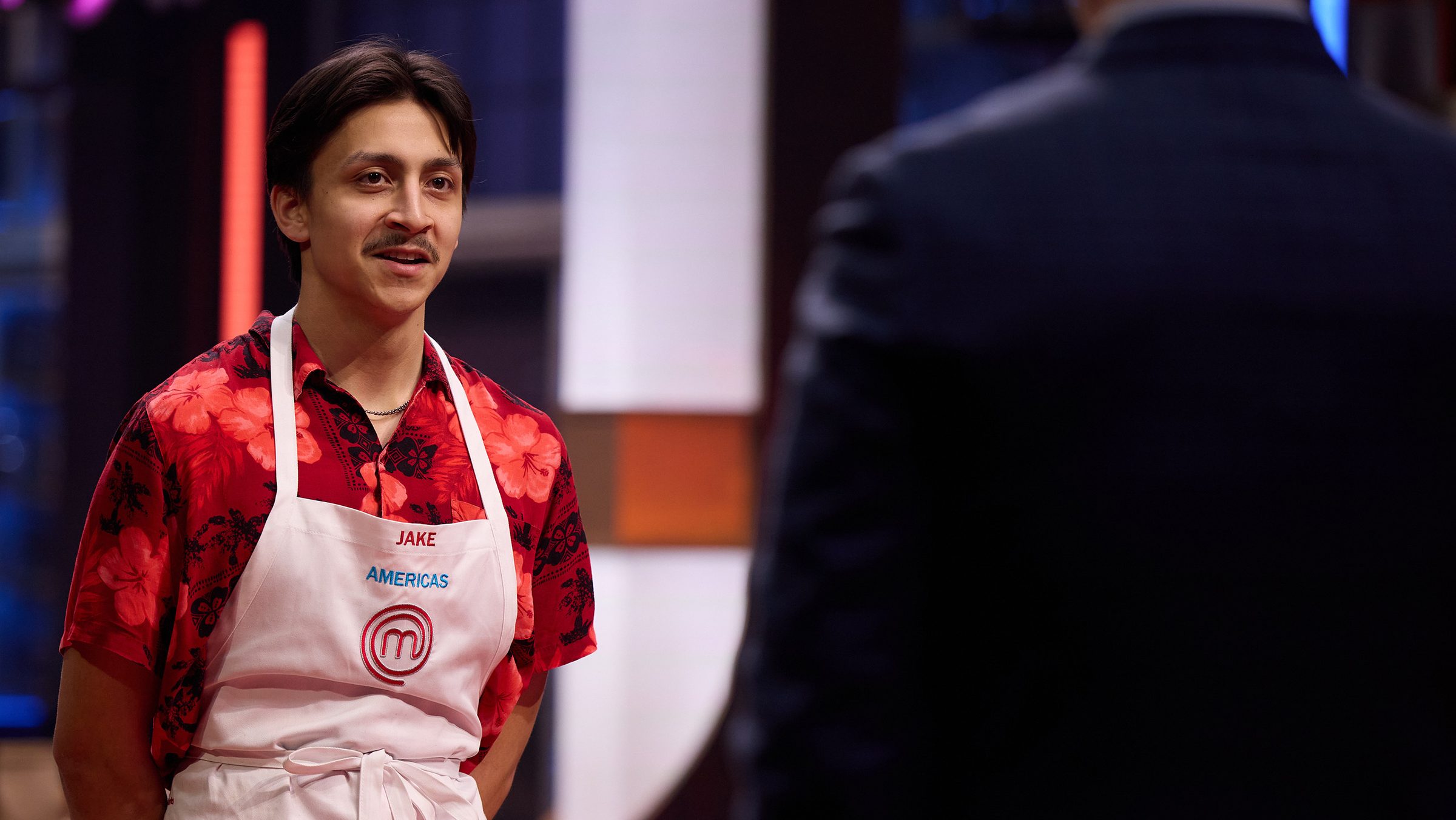 A young man, Jake Kelley, wearing a floral red shirt and a white MasterChef apron, stands and smiles while speaking to another contestant in an intense cooking competition setting.