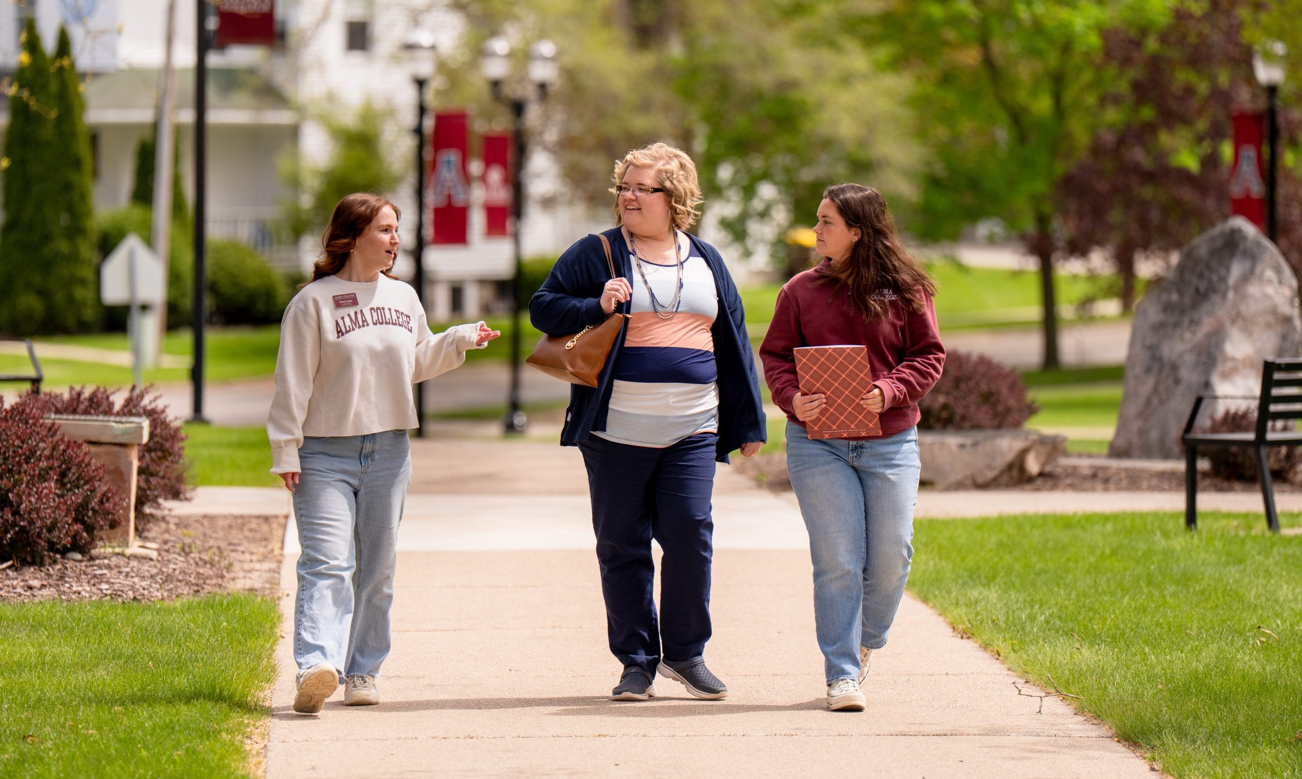Three people are walking and talking on a college campus sidewalk surrounded by green grass and trees. Two hold books or folders, and one wears an Alma College sweatshirt. It’s a bright, sunny day—a perfect moment to connect-and-visit.