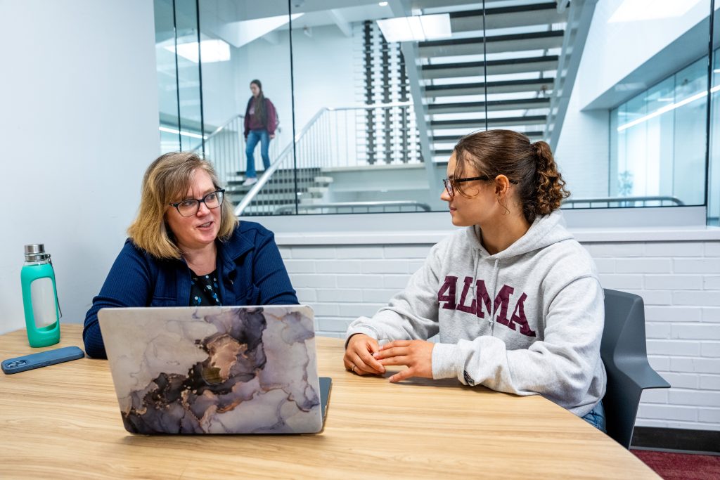 Two women sit at a table with a laptop, talking. One wears glasses and a blue jacket; the other wears glasses and an ALMA hoodie, representing one of the top colleges known for its welcoming community. A person walks upstairs in the background.