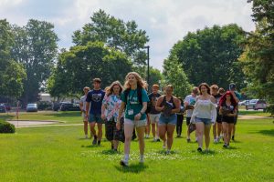 A group of teens walk together on a sunny day across a grassy field, one of them sporting a tartan-101 patterned shirt, surrounded by trees and parked cars in the background.