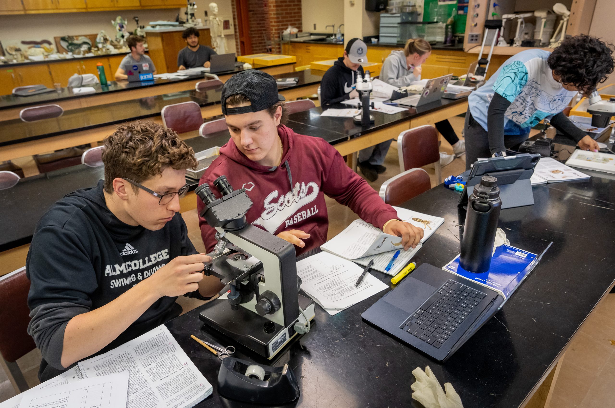 Two students examine a microscope in a busy science classroom while others work at desks with laptops and papers. Models, lab equipment, and diagrams are visible in the background.