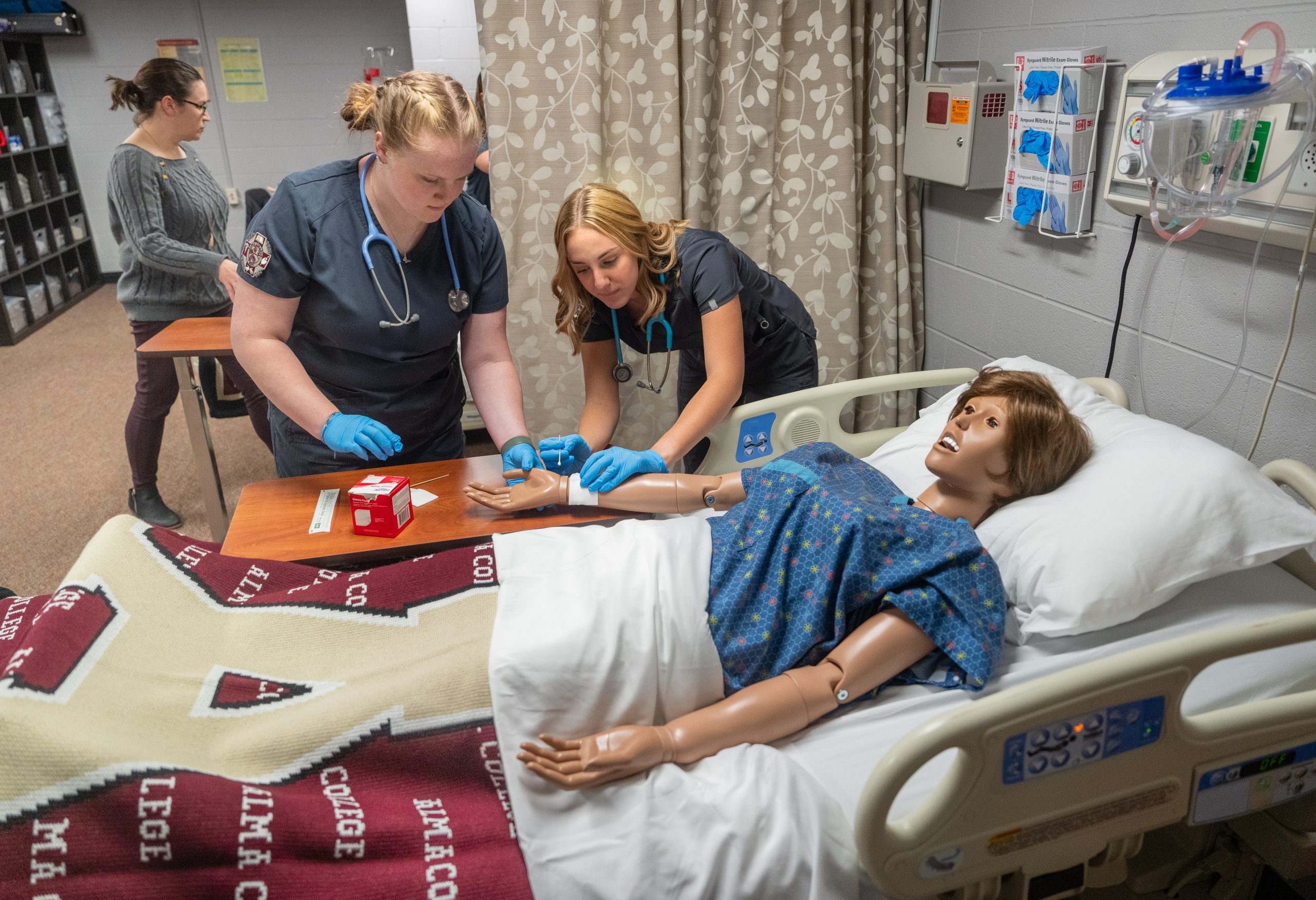 Two nursing students practice medical procedures on a hospital bed mannequin, with one student drawing blood and another assisting, while a third person works in the background. Medical equipment is visible on the wall.