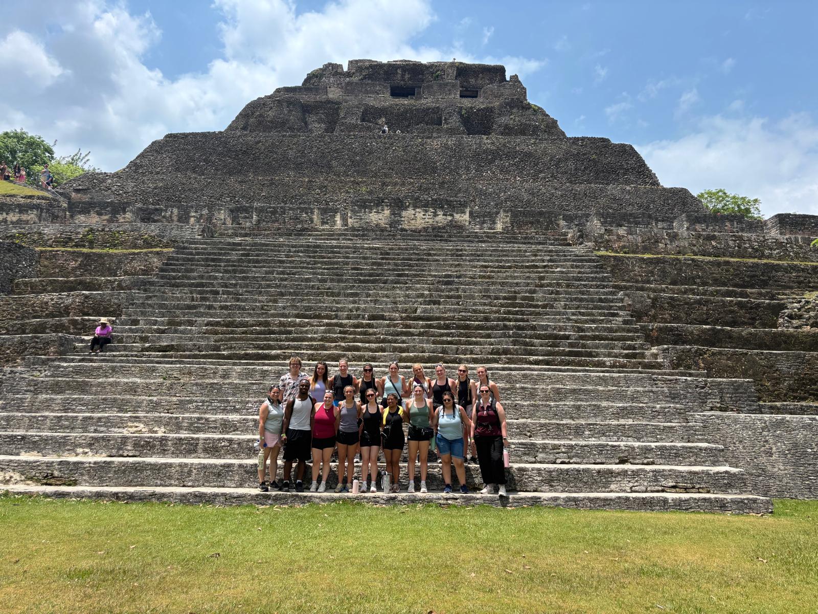 A group of people poses on the steps of an ancient stone pyramid under a partly cloudy sky, with greenery visible at the edges, capturing a memorable moment during their spring-term adventure.