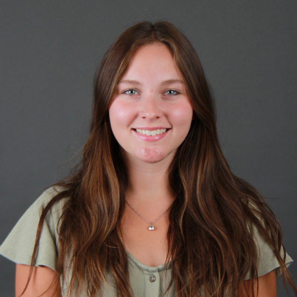 A young woman with long brown hair smiles at the camera. Dilynn Everitt is wearing a light green top and a silver necklace, standing in front of a plain gray background.