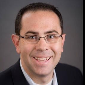 A man with short dark hair, wearing glasses, a dark suit jacket, and a light dress shirt, smiling at the camera against a plain dark background—a professional image suitable for trustees.