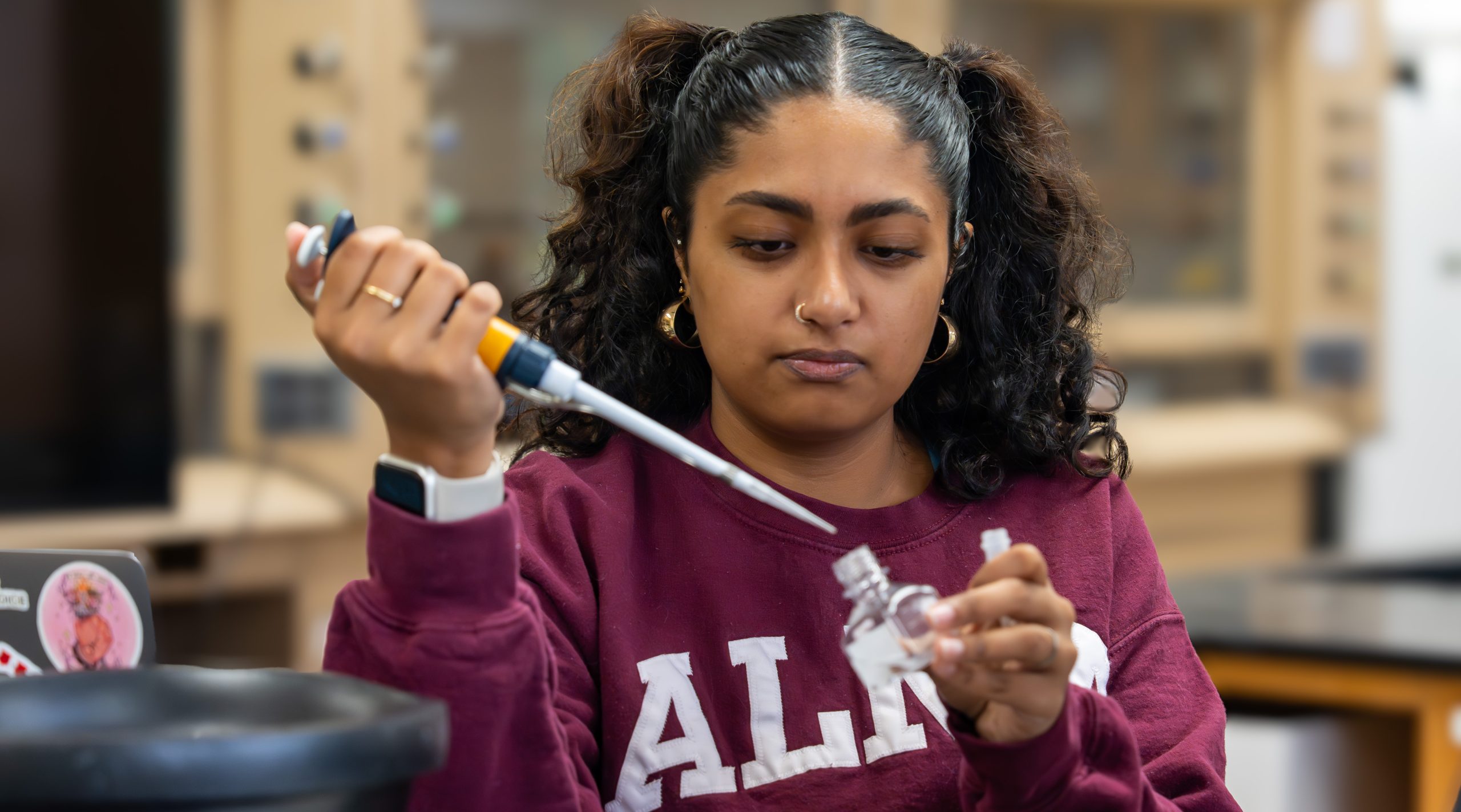 A woman in a maroon sweatshirt uses a pipette to transfer liquid into a small glass bottle in a laboratory setting, exemplifying hands-on research opportunities available through Life Sciences at Alma College.