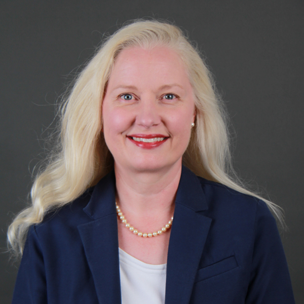 Kerrissa Silverthorne, a woman with long blonde hair, wearing a navy blazer, white top, pearl necklace, and red lipstick, smiles in front of a plain dark gray background.