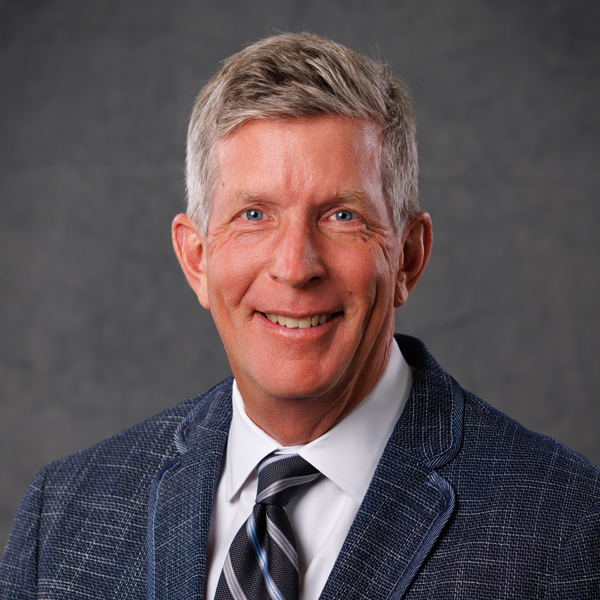 A smiling older man with short gray hair, wearing a blue textured blazer, white shirt, and striped tie, poses in front of a plain gray background—reflecting the professionalism often associated with trustees.
