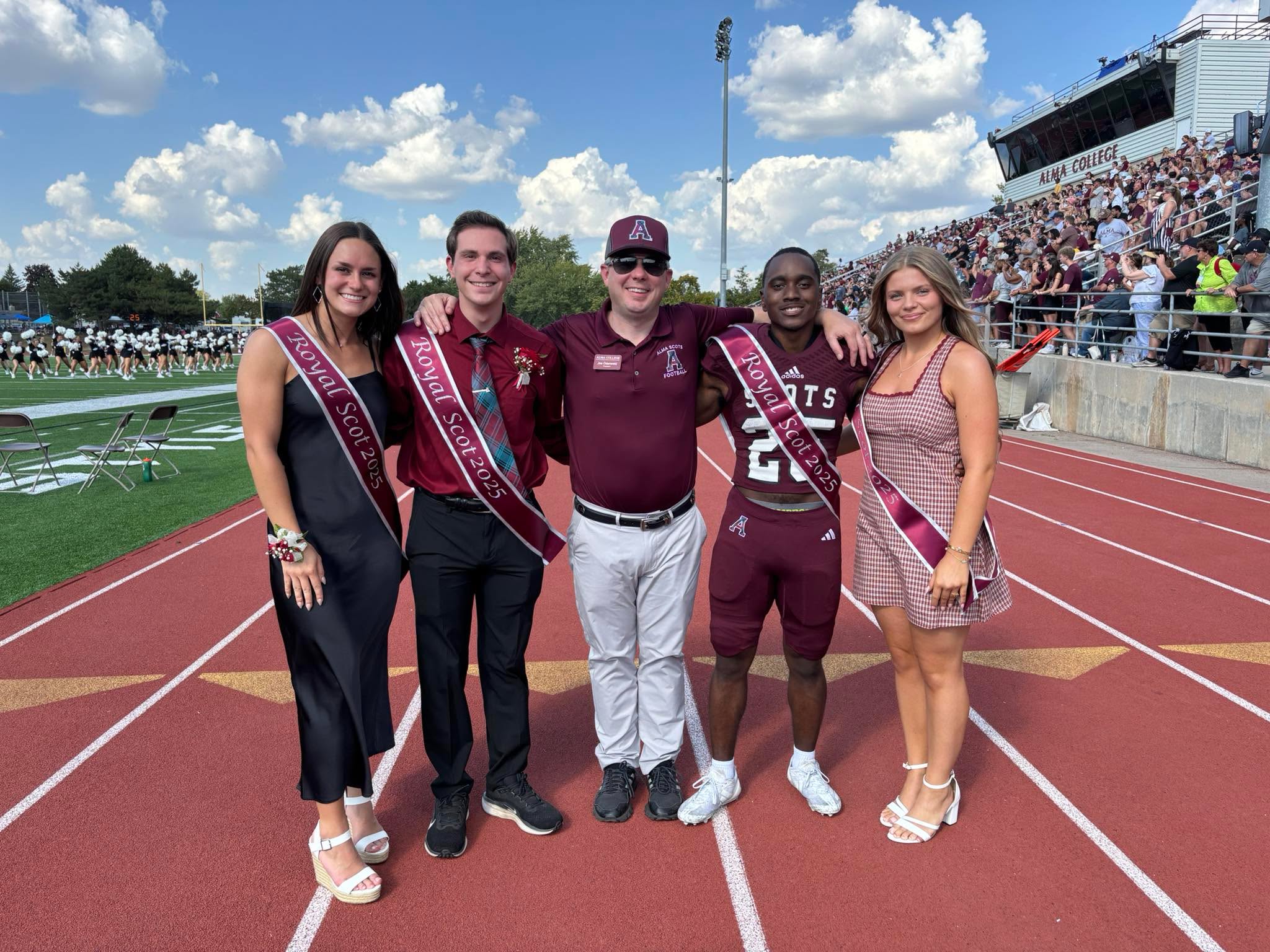 Five people wearing sashes pose together on a football field sideline during a game, capturing the spirit of life at Alma College, with a crowd in the stadium and a marching band performing in the background on a sunny day.