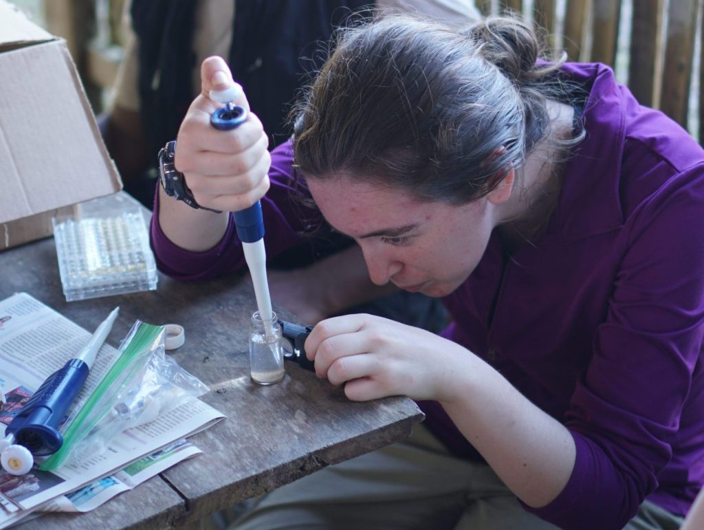 A person uses a pipette at a wooden table covered with lab supplies while another person watches, showcasing hands-on learning in Life Sciences at Alma College. They are working outdoors near a bamboo fence.