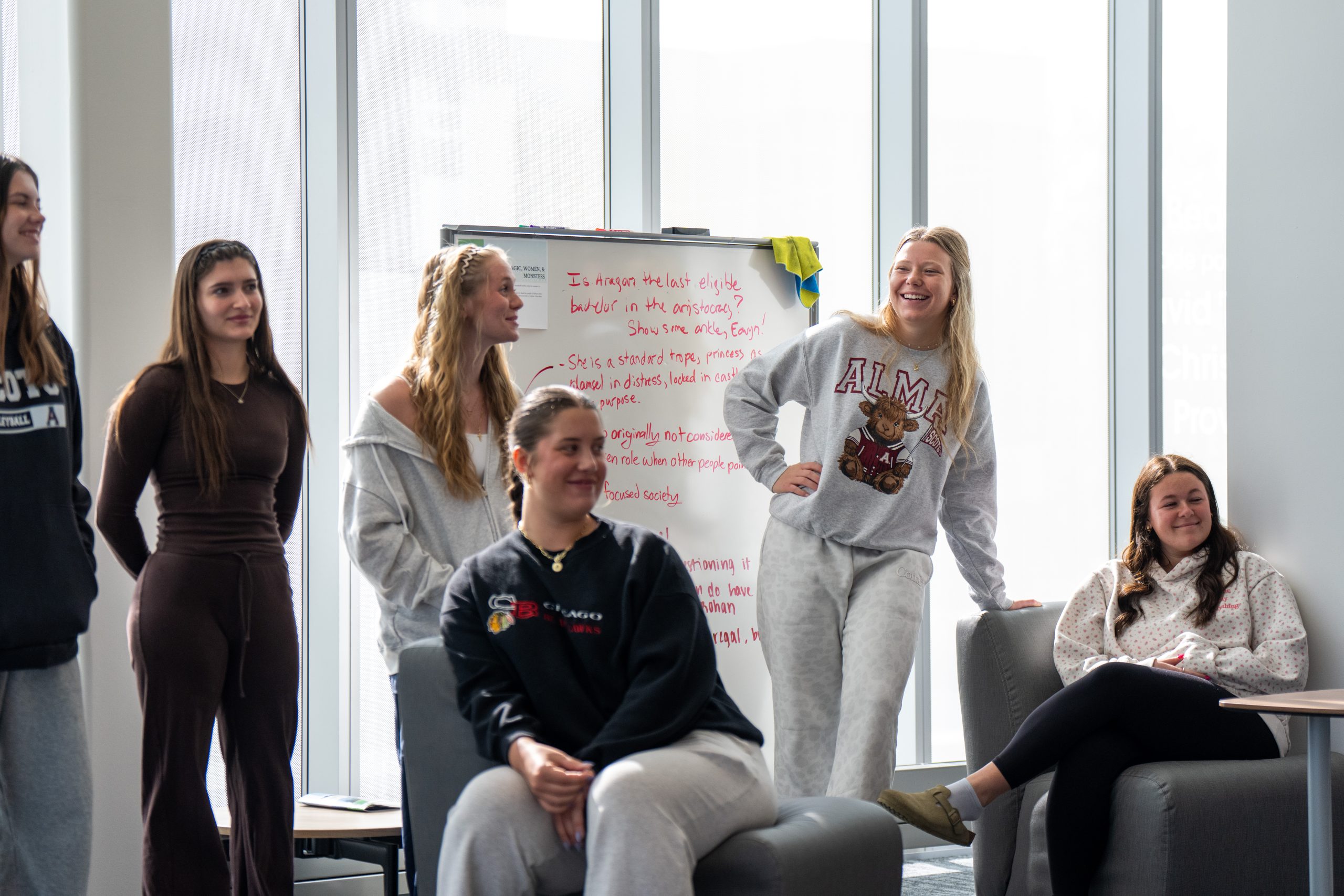 Six young women are gathered in a bright room, some sitting and others standing, smiling and talking—capturing a lively moment of life at Alma College.