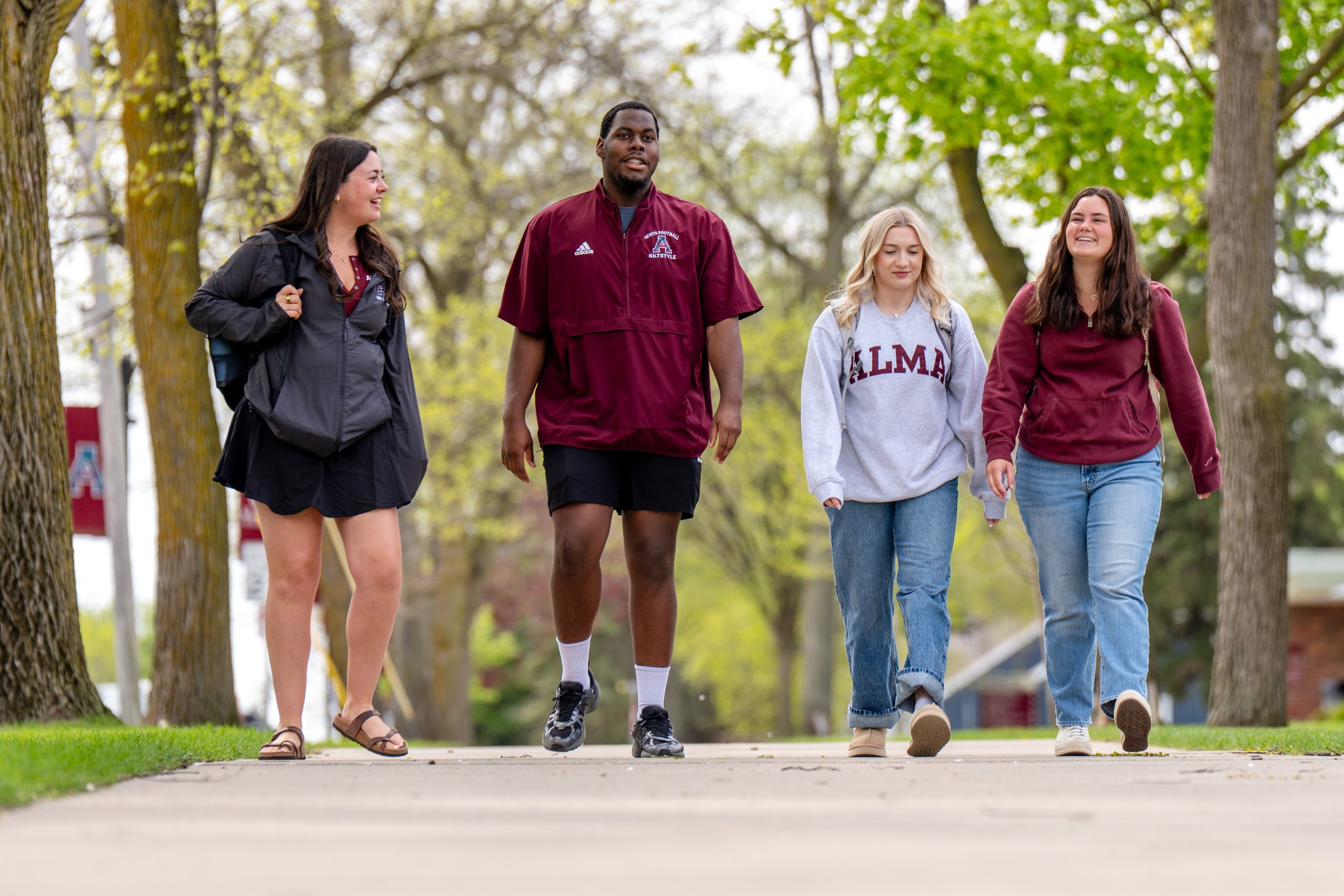 Four college students, three women and one man, walk outside on a tree-lined sidewalk, smiling and talking together. Dressed in casual clothes—some sporting Alma College logos—they capture the friendly spirit of life at Alma College.