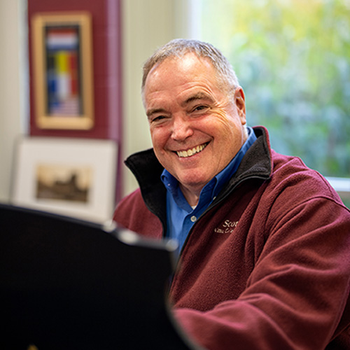 Smiling man in a maroon fleece sits at a desk or piano indoors, with framed art and photos on the wall behind him.