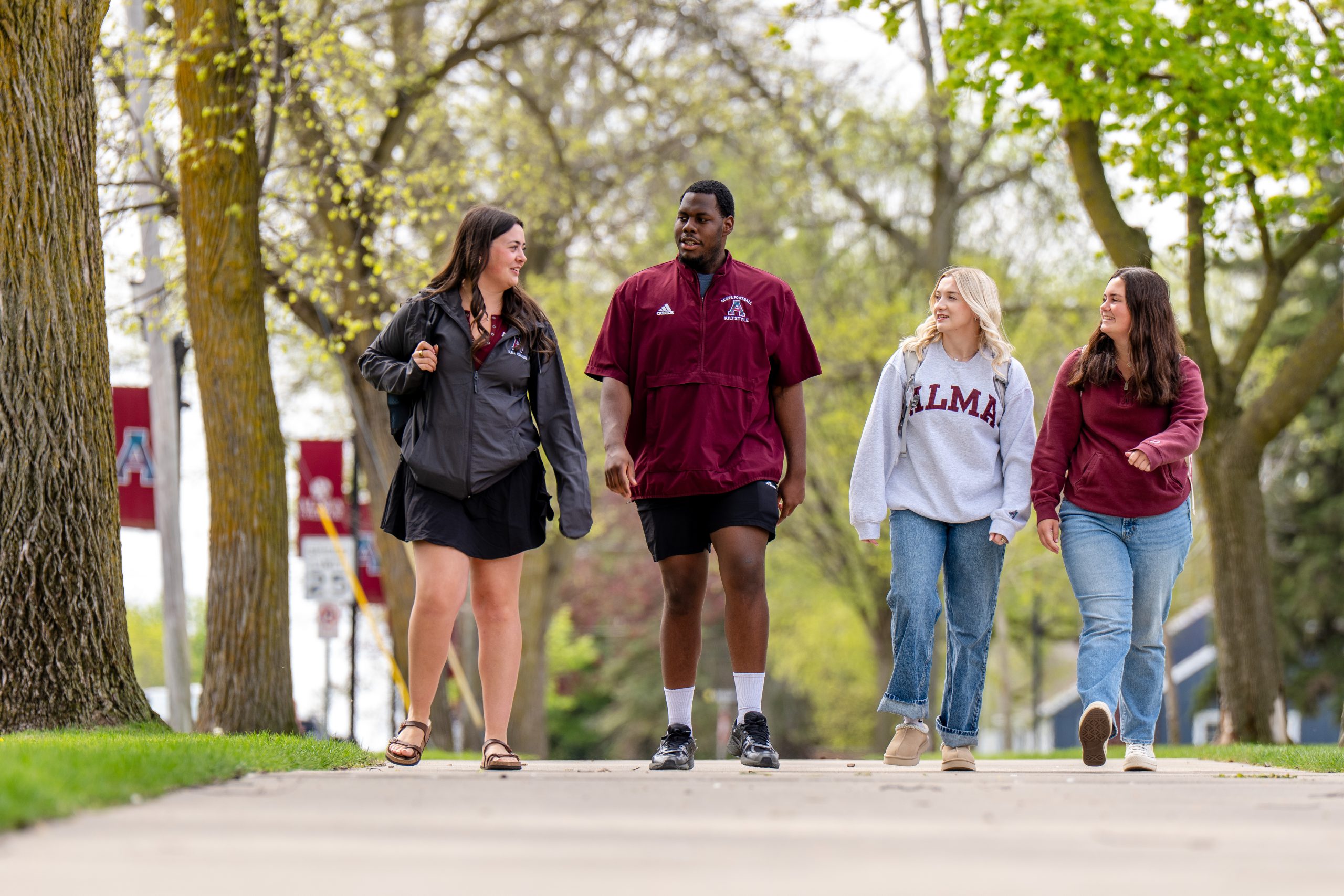 Four college students walk together on a tree-lined campus sidewalk, smiling and talking about the college application process. Two wear Alma College clothing, while spring leaves and campus signs are visible in the background.