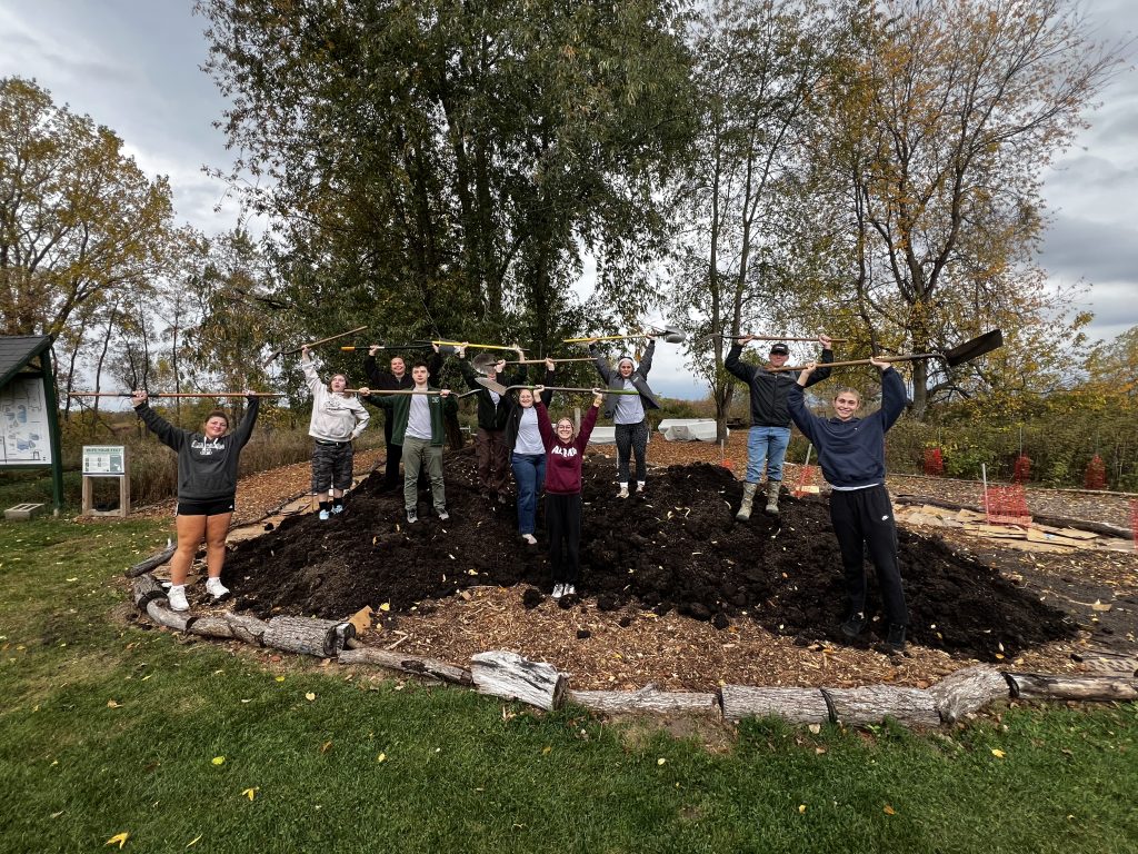 A group of people stand on a mulch mound outdoors, holding shovels above their heads and smiling at the camera for a 3CE News feature. Trees with autumn leaves and cloudy skies are in the background.