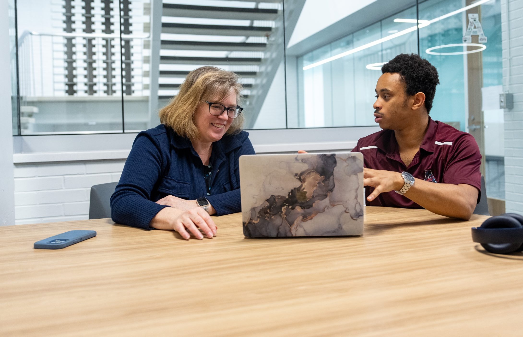 Two people sit at a table in the registrar’s office, smiling and talking while looking at a laptop. They are in a modern office with glass walls. Headphones, a phone, and a water bottle are on the table.