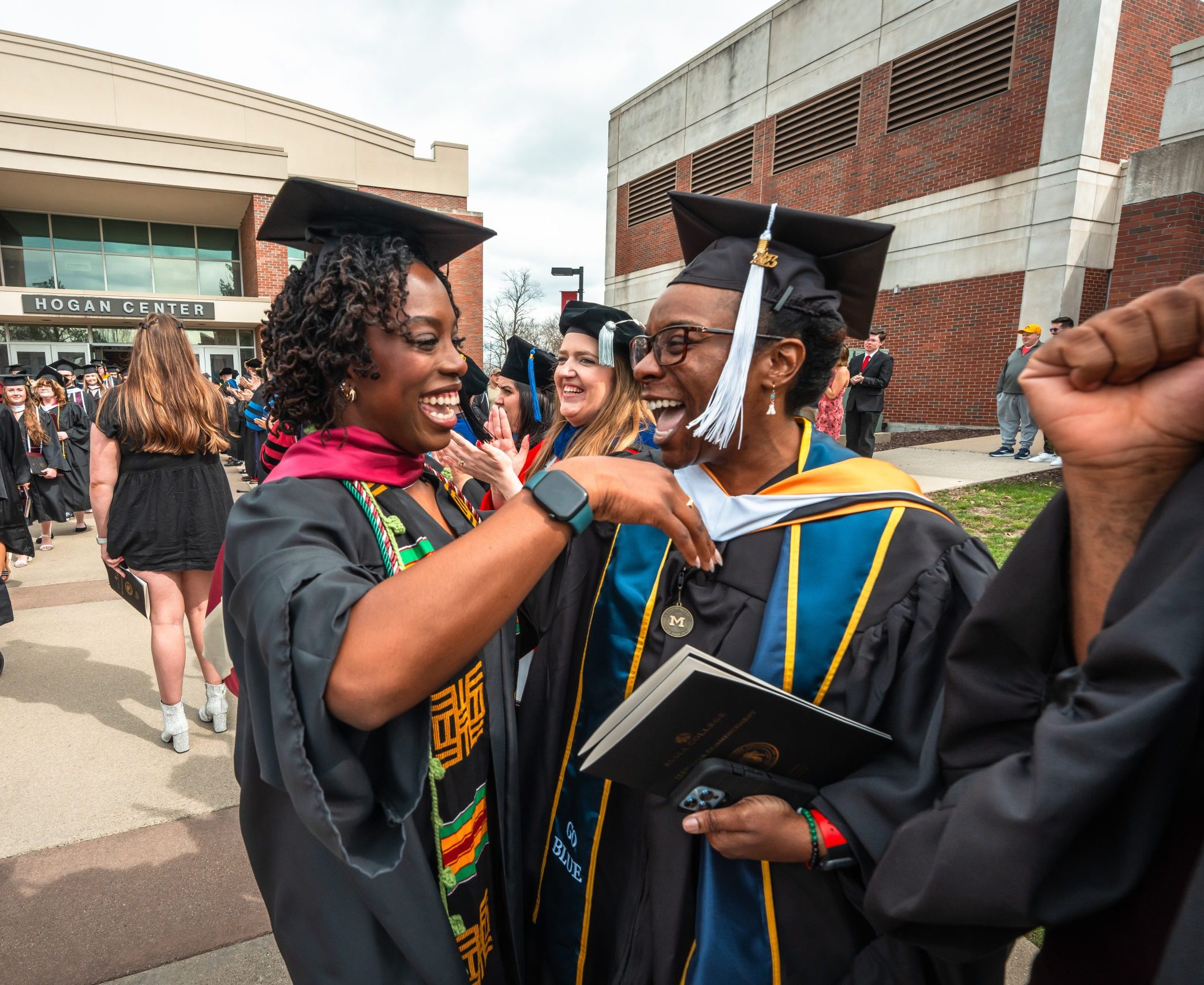 Two graduates in caps and gowns smile and celebrate together outside a campus building during commencement at Alma College, surrounded by fellow graduates and people in the background.