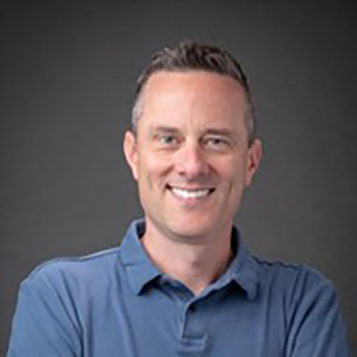 Smiling man with short brown hair wearing a blue collared shirt stands with arms crossed against a plain dark background.