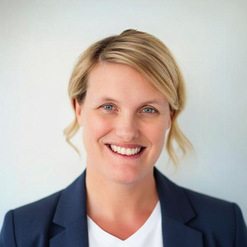 Smiling woman with blond hair, wearing a navy blazer and white top, standing in front of a plain light background.