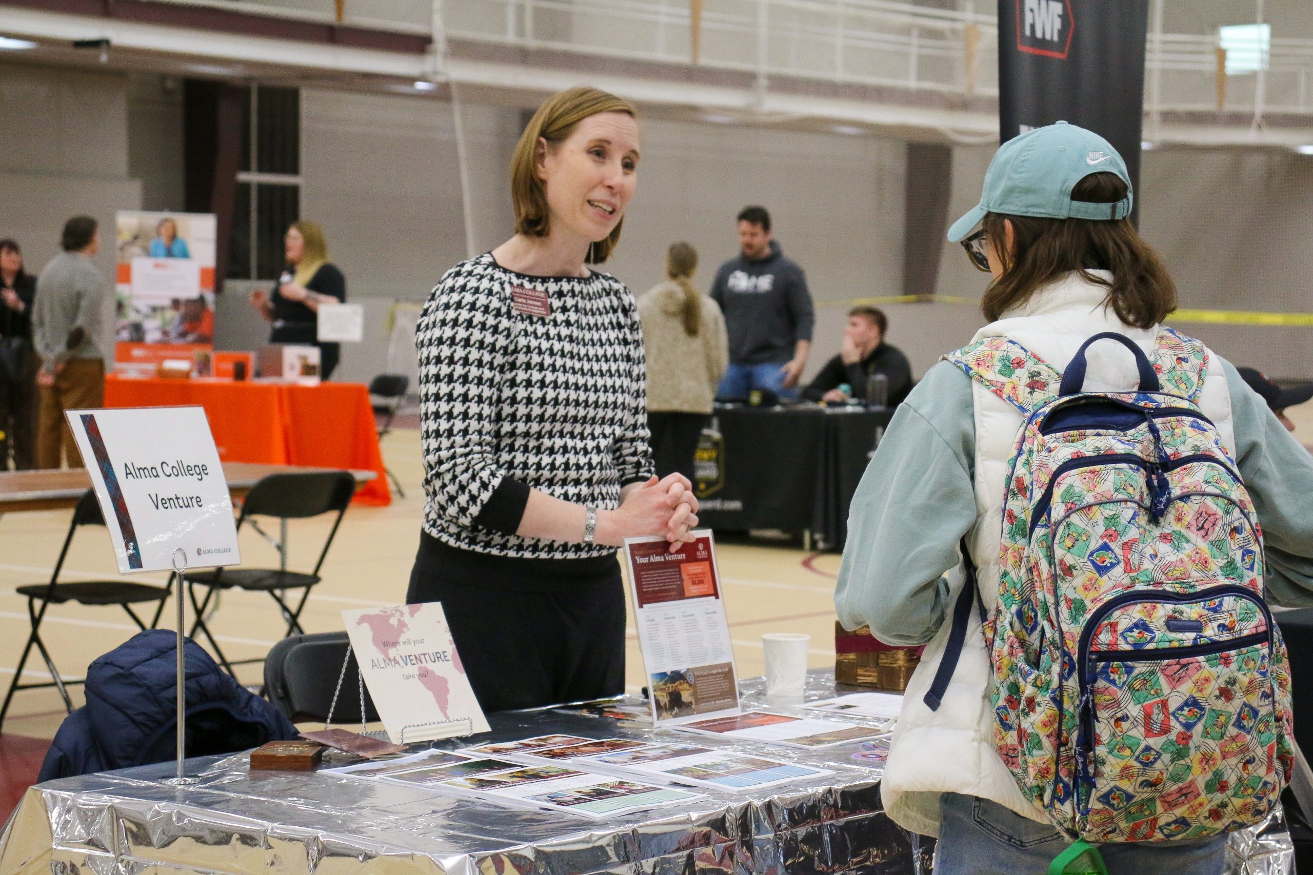 A woman stands behind a table with Alma College Venture materials, discussing personal development opportunities with a student wearing a cap and patterned backpack at an indoor college event. Other tables and people are visible in the background.