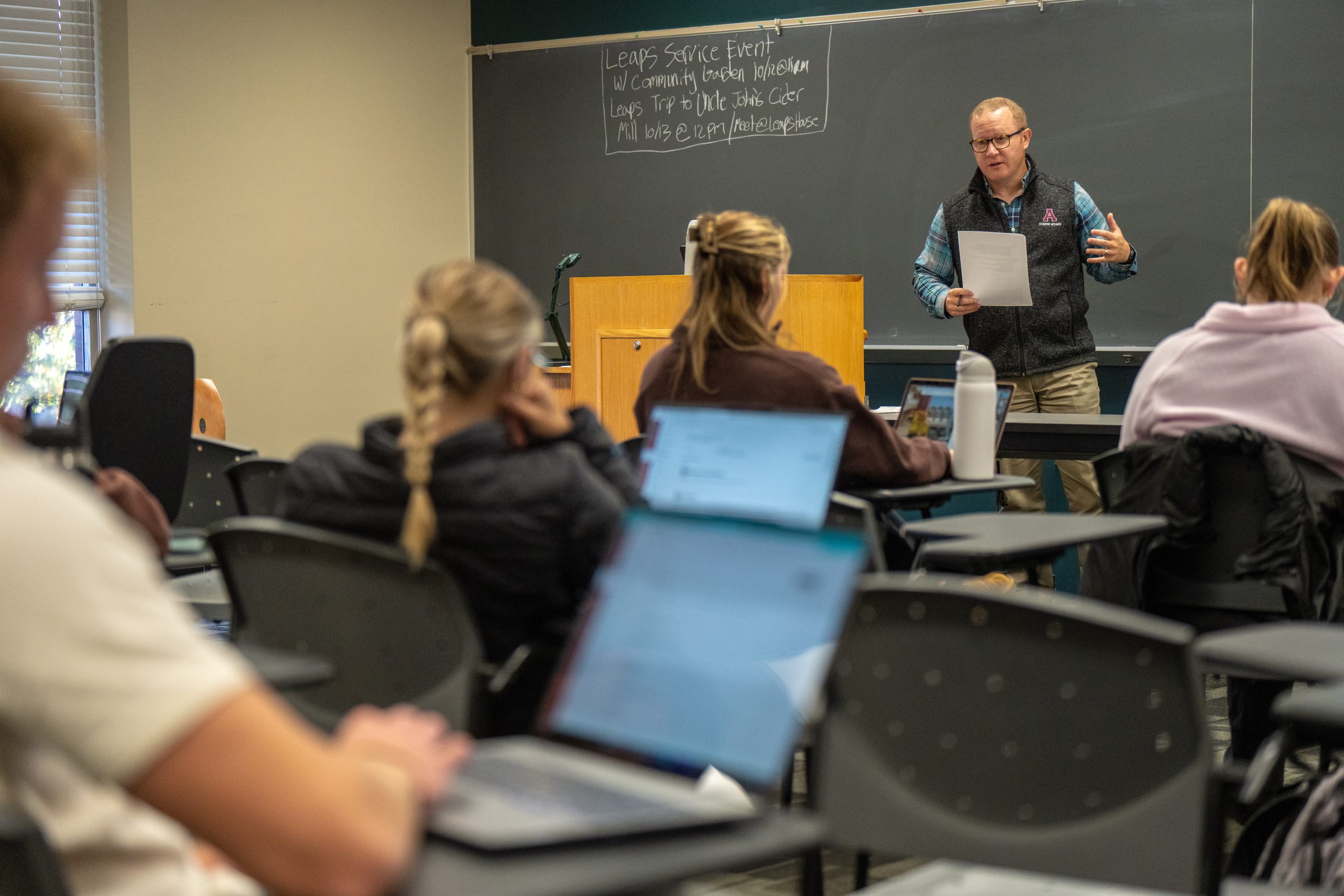 A teacher stands at the front of a classroom, speaking to students who are seated at desks, some with laptops open as part of their learning. Notes are written on the chalkboard behind the teacher.