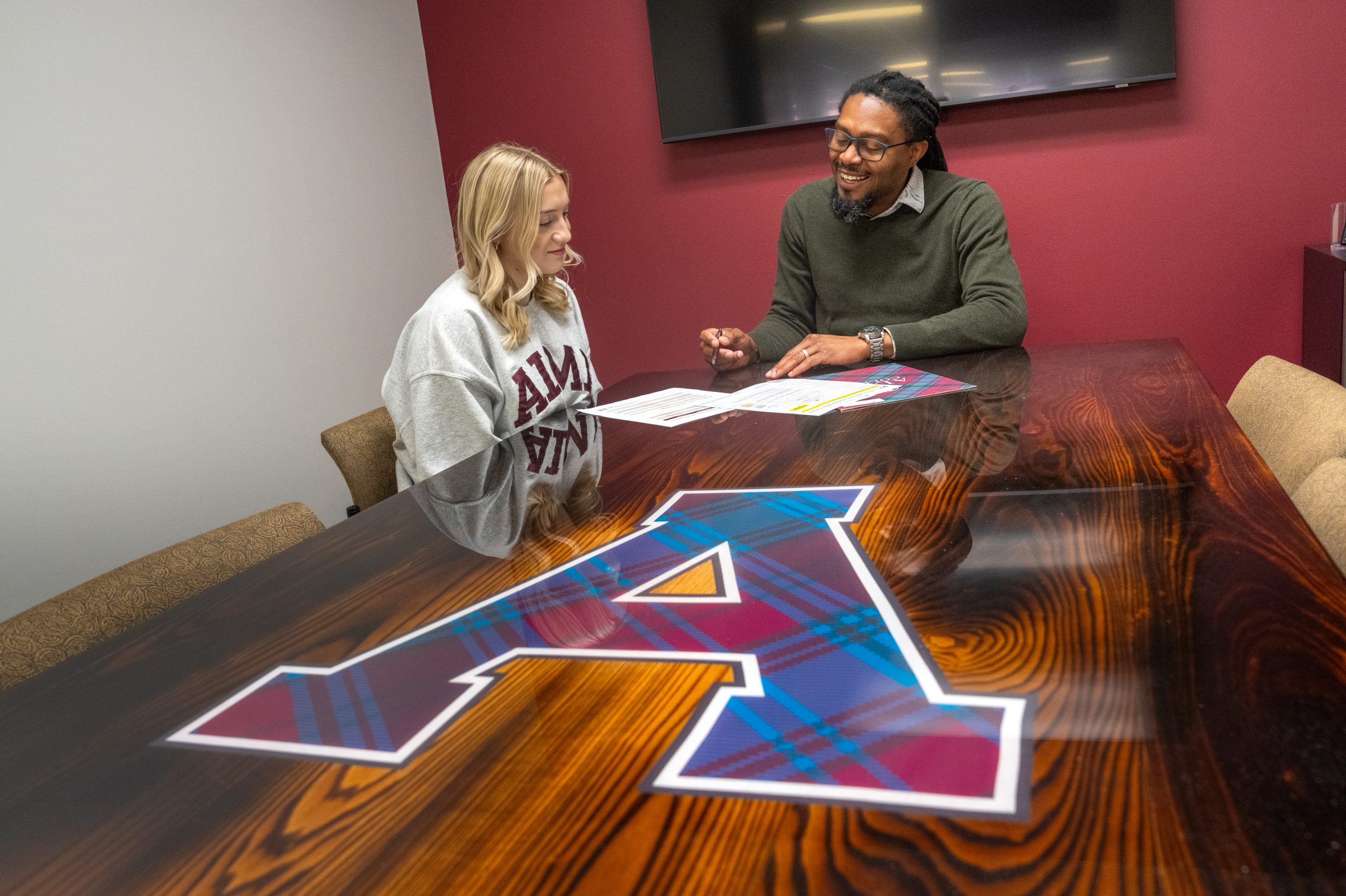 Two people sit at a polished table with a large letter A decal, discussing financial-aid papers. One person wears an A&M sweatshirt. The setting appears to be a casual office or meeting room with maroon and white walls.