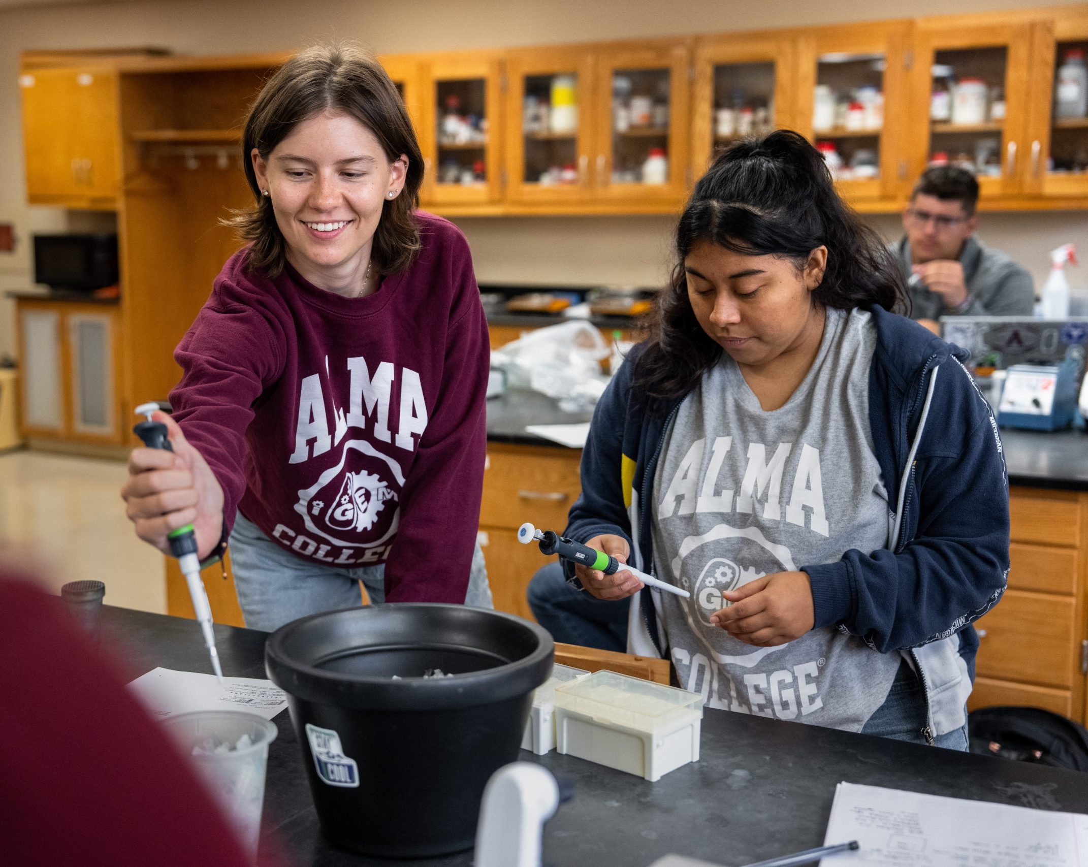 Two students in Alma College shirts are experimenting with pipettes in a science lab. One student smiles and points while the other focuses on their task, showcasing their enthusiasm as IGem students. Lab equipment and shelves with supplies are visible in the background.