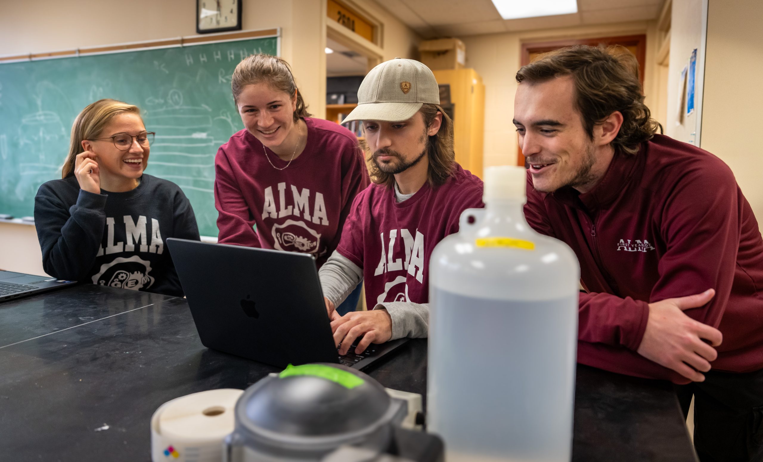 Four college students in Alma College shirts gather around a laptop, smiling and working together in a classroom with a chalkboard and science equipment visible, as they collaborate on their entry for the international genetically engineered machine competition.