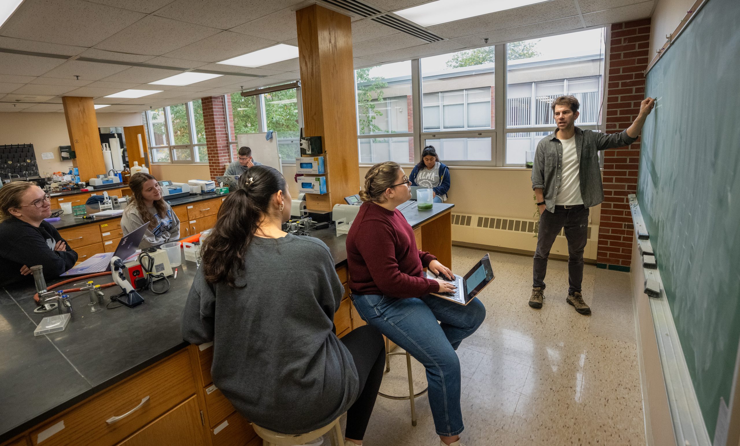 A young man stands at a chalkboard explaining to five people seated around a lab classroom. Some listen, some take notes, and lab equipment is visible on the counters from an International Genetically Engineered Machine (iGEM) project.