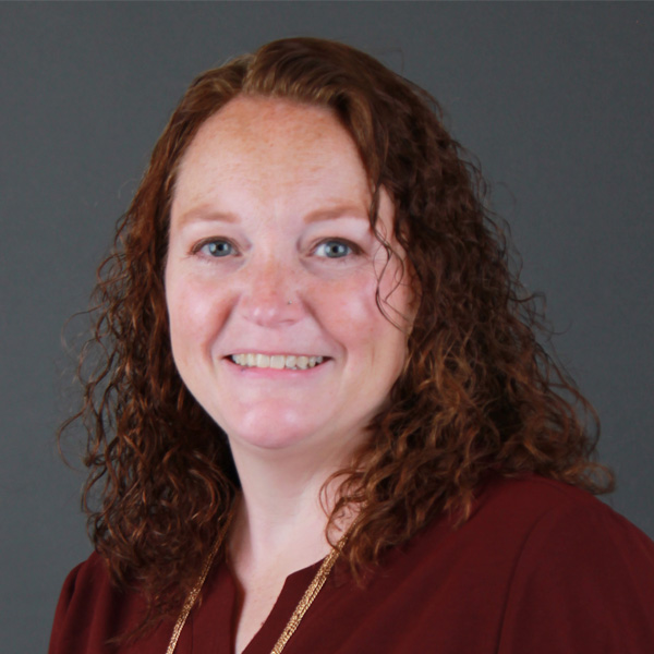 Jennifer Wentworth, a woman with shoulder-length curly red hair, wears a burgundy top and a gold necklace, smiling in front of a plain dark gray background.