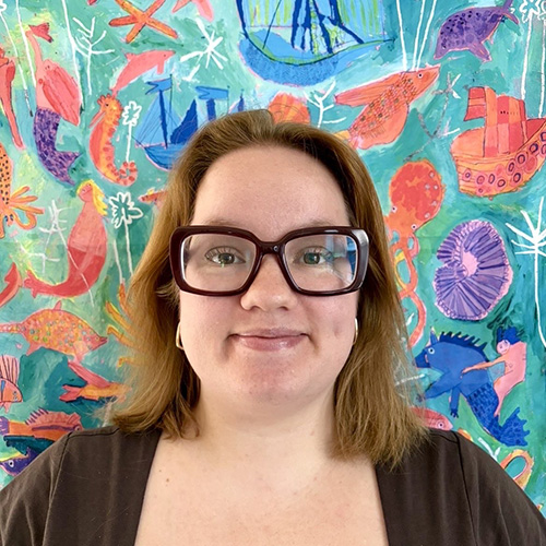 A person with shoulder-length brown hair and large glasses smiles in front of a colorful, sea-themed mural with fish, ships, and sea creatures on a blue-green background.
