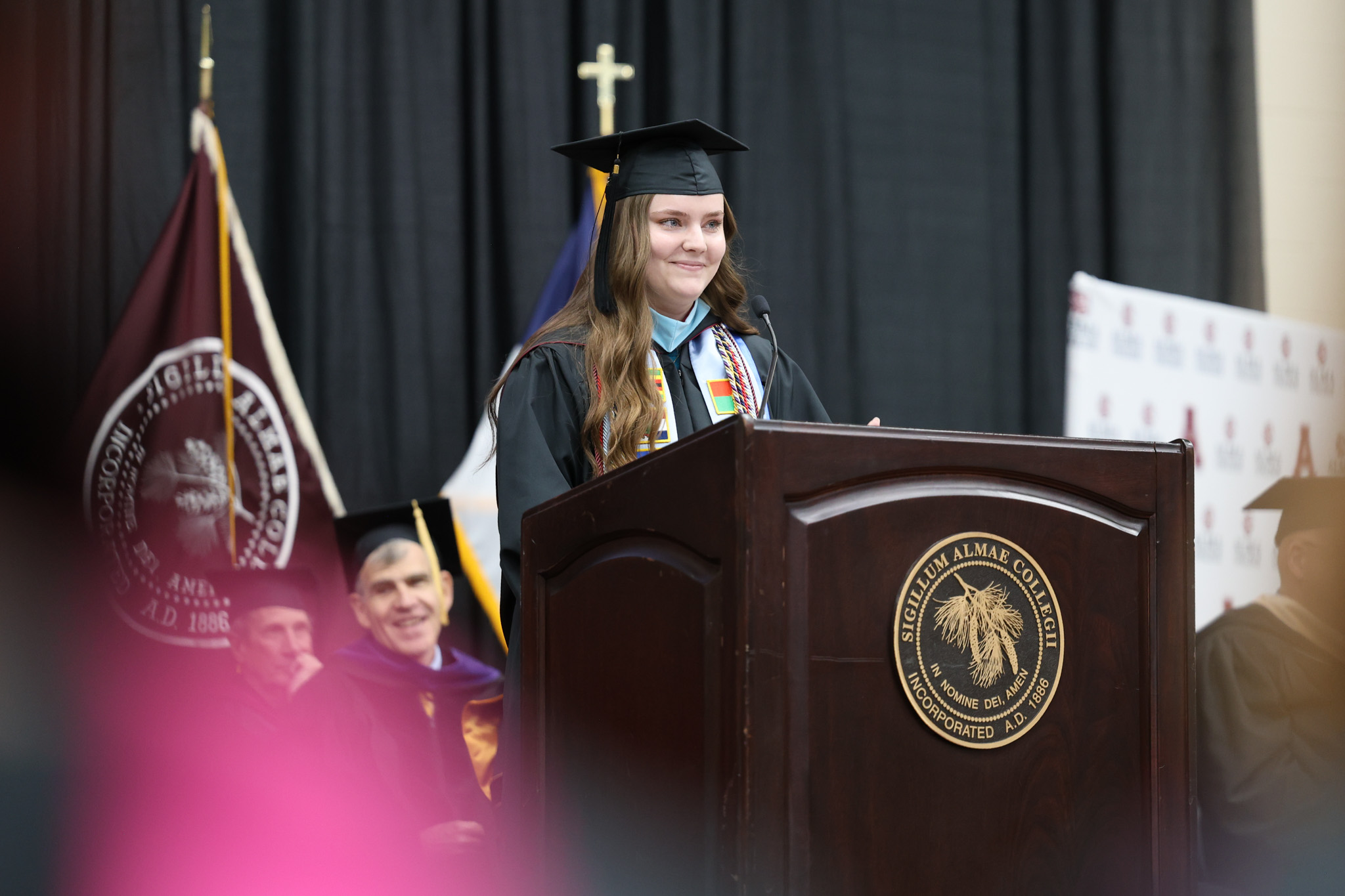 A graduate in cap and gown stands at a podium with a college seal, speaking at a commencement at Alma College ceremony. People in academic regalia are seated behind her, and flags are visible in the background.