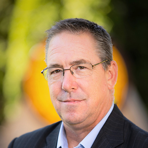 A middle-aged man with short brown hair, glasses, and a slight smile wears a dark suit and light blue shirt, standing outdoors with a blurred green and yellow background.