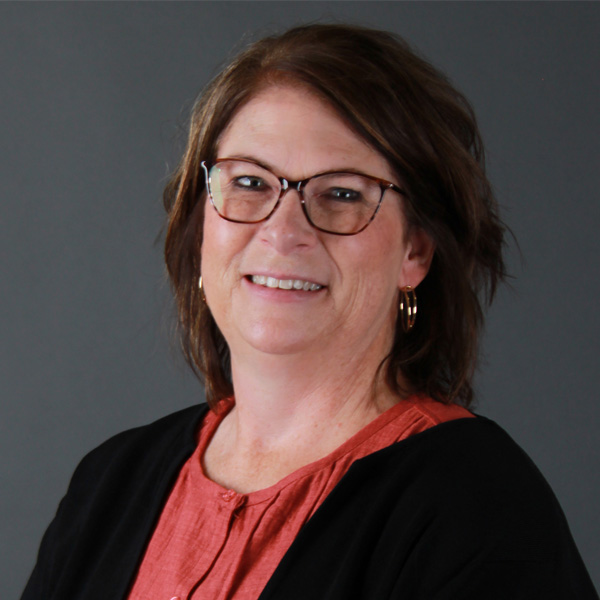 Sandee Gadde, a woman with shoulder-length brown hair, wearing glasses, a coral blouse, a black cardigan, and gold hoop earrings, smiles in front of a plain gray background.