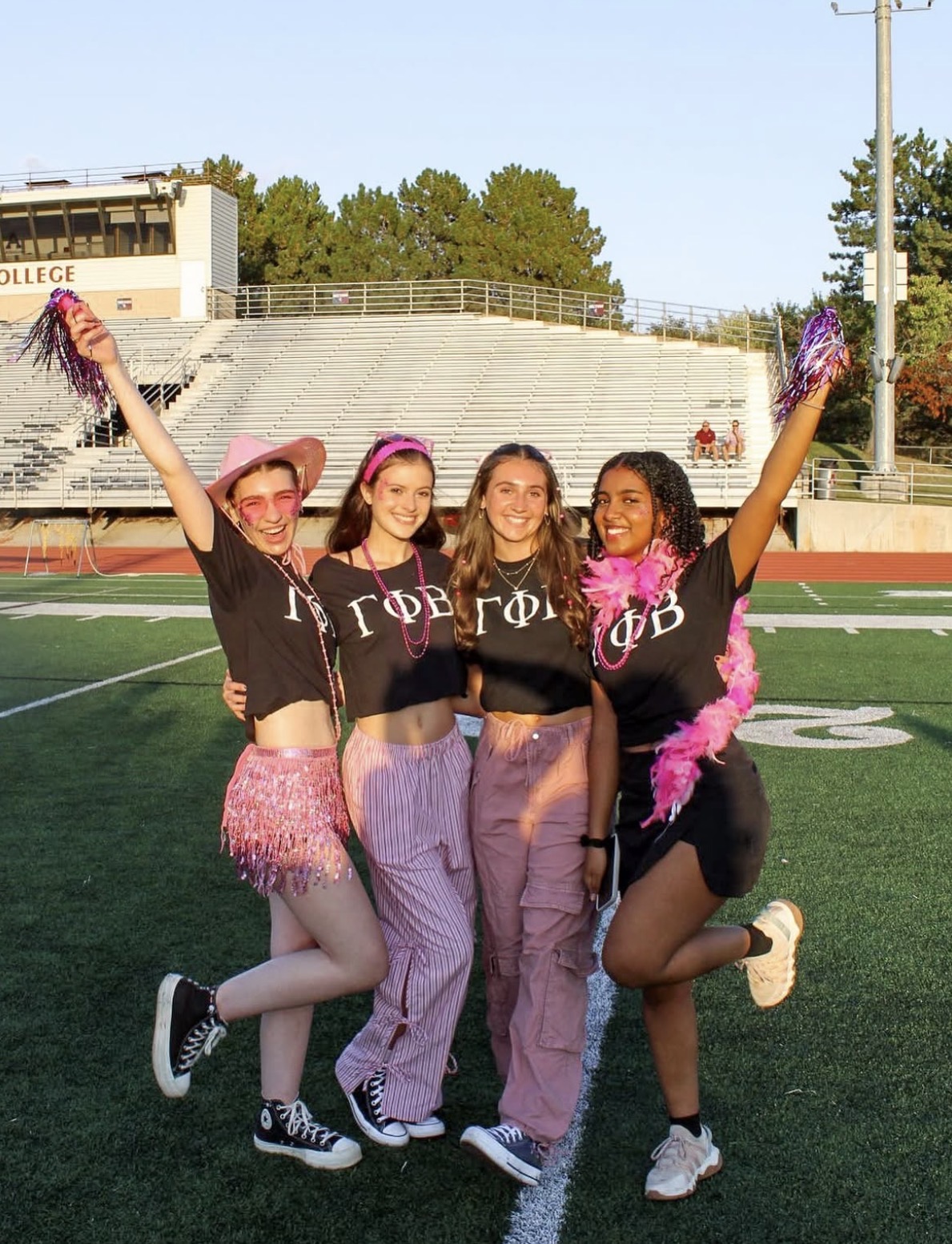 Four young women in matching black shirts and pink accessories stand on a football field, smiling and posing together. This vibrant moment captures the spirit of life at Alma College, with bleachers and trees in the background under a clear sky.