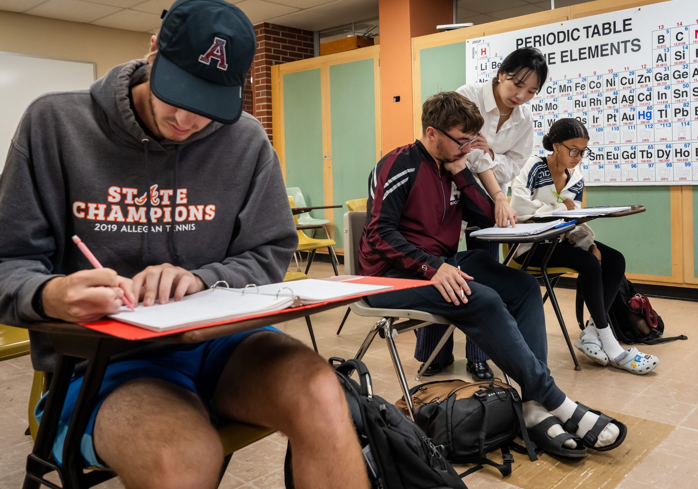 Three students sit at desks in a classroom with a periodic table on the wall. One student gets help from a professor while others focus on their work. The class looks casual and studious.