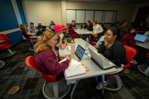 Students sit in groups at desks with laptops and notebooks in a classroom, working together on assignments as part of a first year seminar. The room has red chairs, gray tables, and a mix of natural and artificial light.