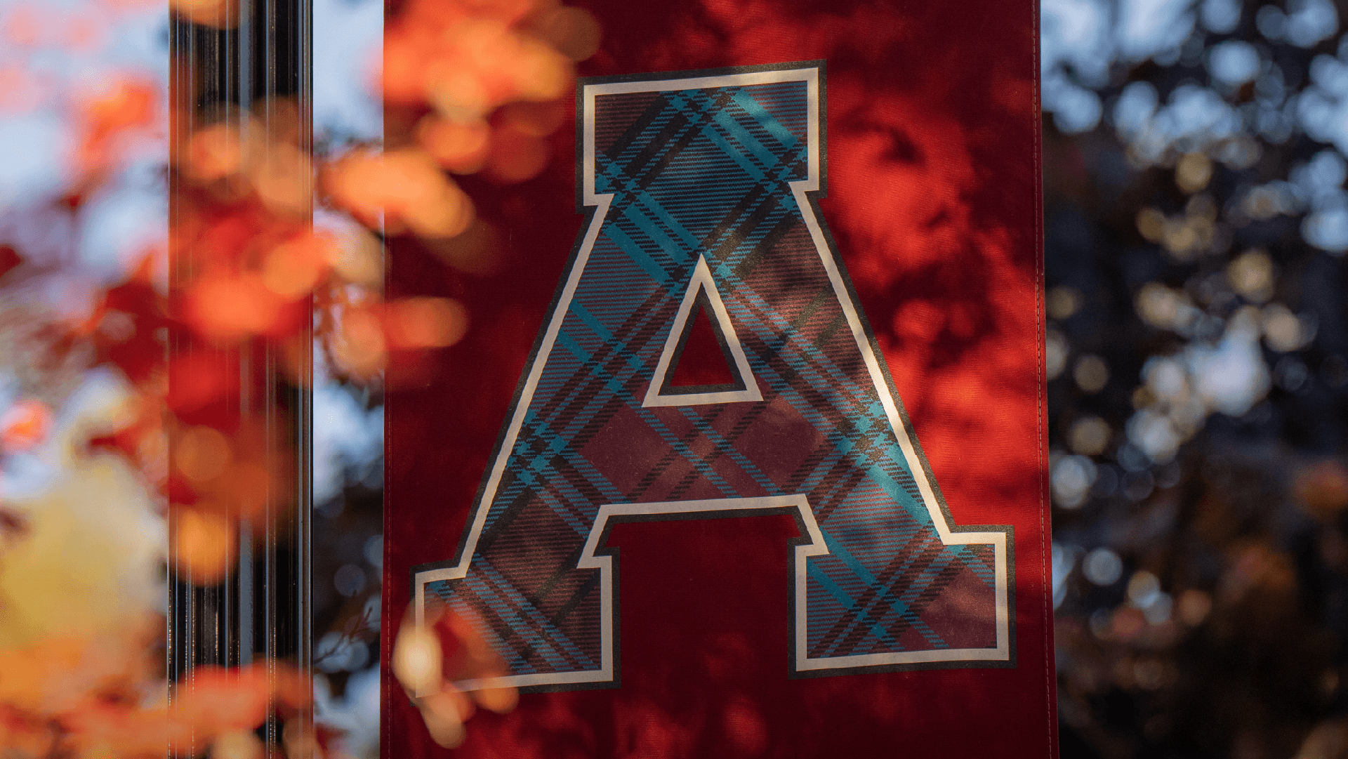 A large letter A with a blue plaid pattern, representing the Alma College Tartan Award, is displayed on a red banner, surrounded by blurred autumn leaves and branches in the background.