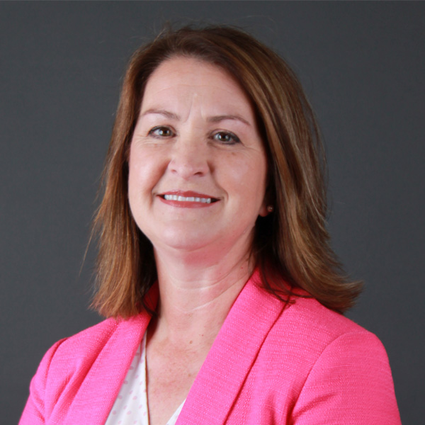 Wendy Judge, a woman with shoulder-length brown hair, is smiling while wearing a pink blazer over a white shirt, set against a gray background.