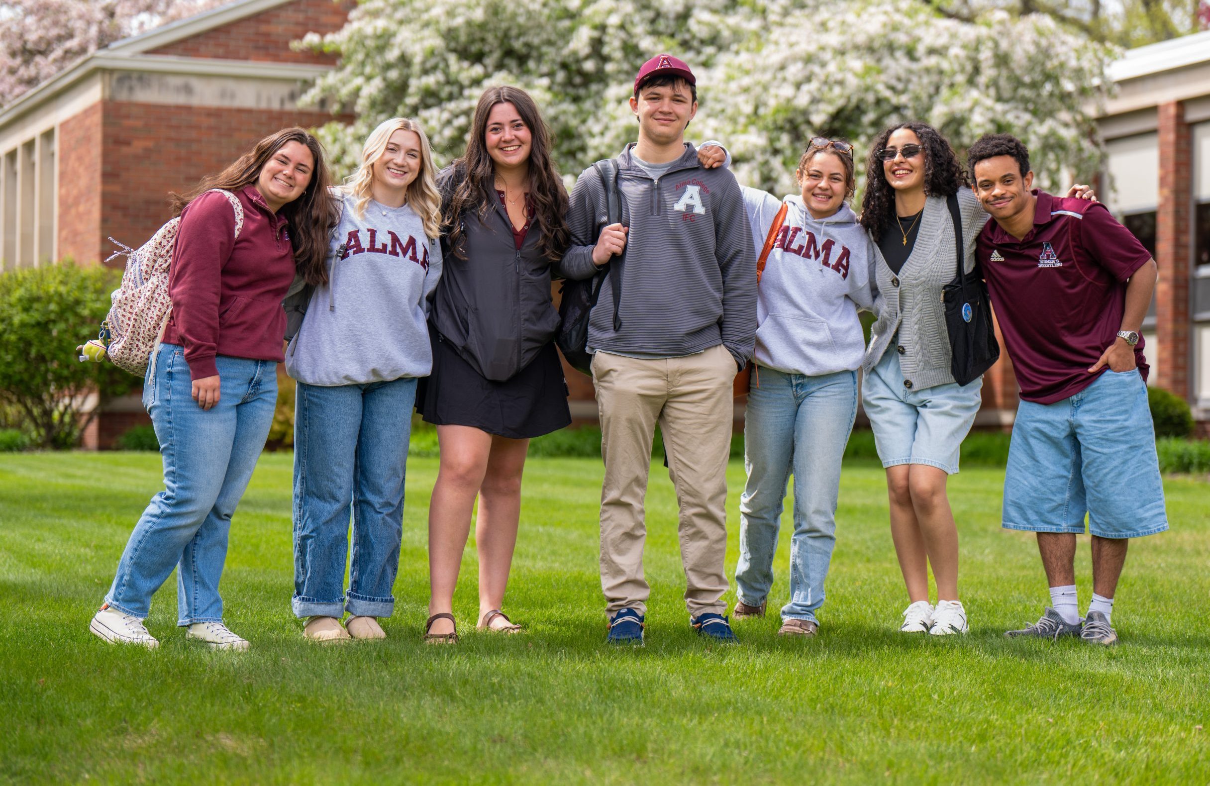 Seven students stand together smiling on a grassy lawn in front of a brick building with blooming trees, wearing casual clothes and some with ALMA sweatshirts, inspiring others to apply and become part of this vibrant community.