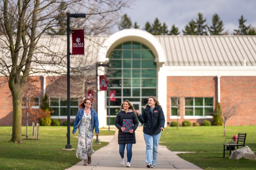 Three people walk and talk on a sidewalk in front of a brick campus building with large windows. The building, adorned with Alma College banners, is surrounded by grass and trees—a perfect spot to connect-and-visit while experiencing campus life.