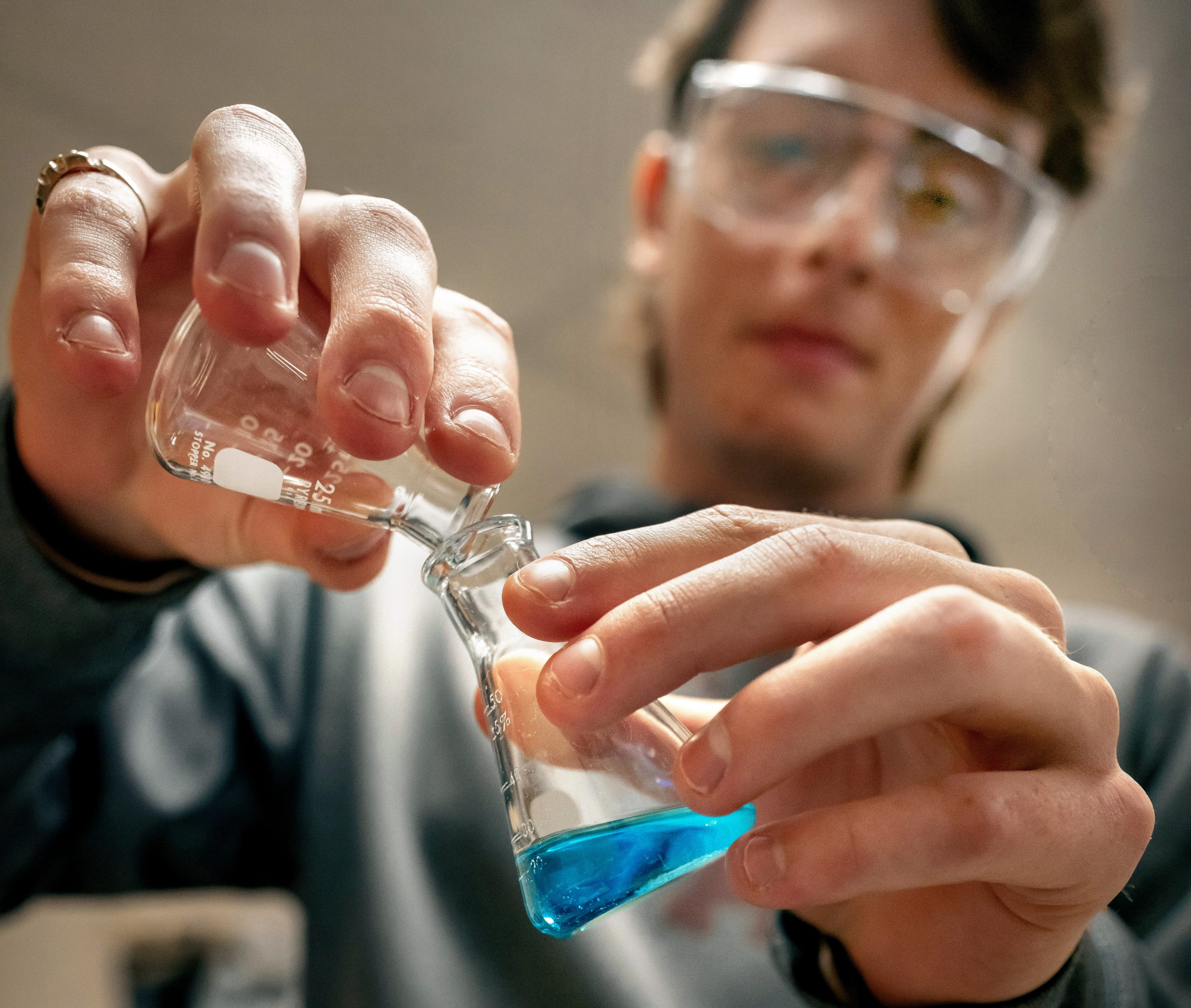 A person wearing safety goggles pours a blue liquid from one glass container to another in a laboratory setting, demonstrating hands-on learning as part of the Chemistry Pathway at Montcalm Community College.