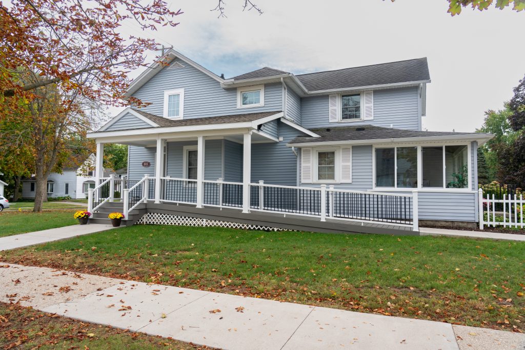 A light blue two-story house with white trim, a large front porch, accessibility ramp, and well-kept lawn offers comfortable overnight-accommodations. Trees with autumn leaves are around the property.