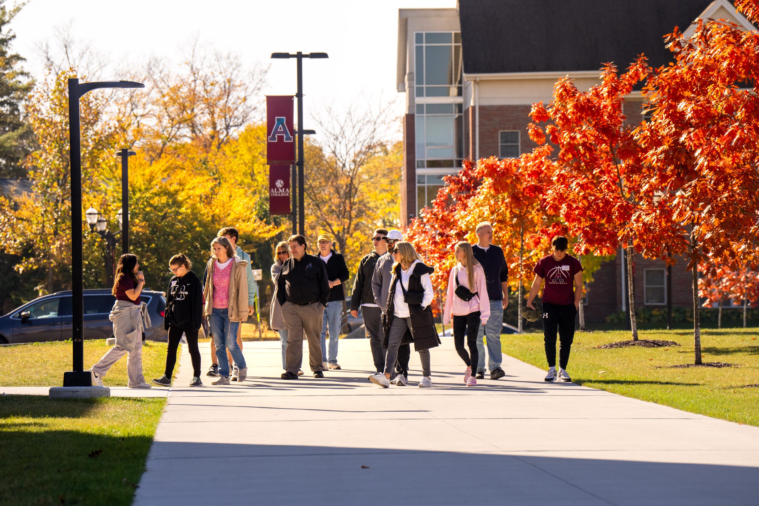 A group of people walk on a sunny campus sidewalk lined with autumn trees and a red banner, taking the opportunity to connect-and-visit as they enjoy the vibrant surroundings. A brick building is visible in the background.