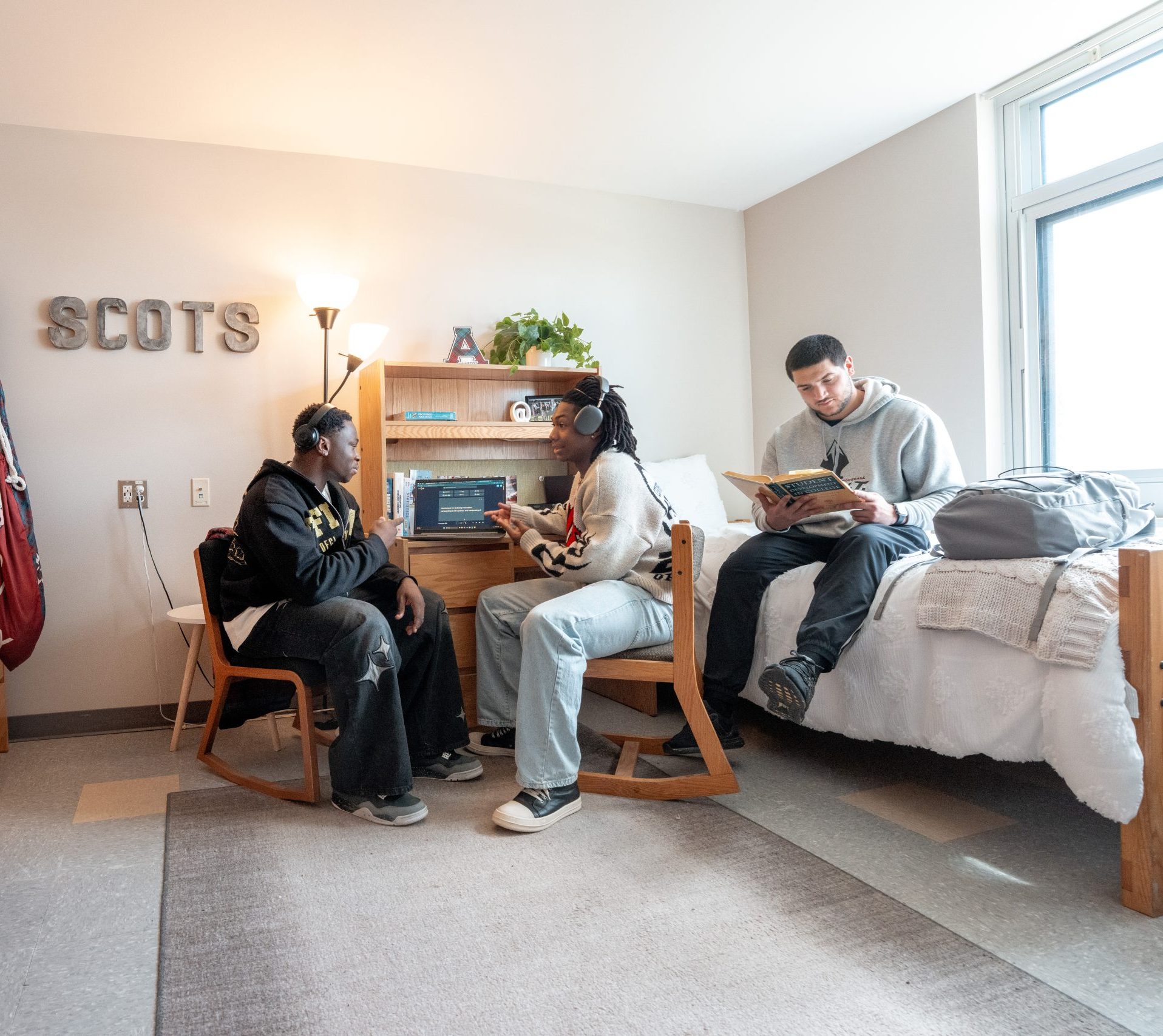 Three students hang out in a college dorm room; two sit talking at a desk, discussing their assignments, while one sits on a bed reading a book. The bright space, decorated with SCOTS letters on the wall, creates an inviting atmosphere for students studying together.