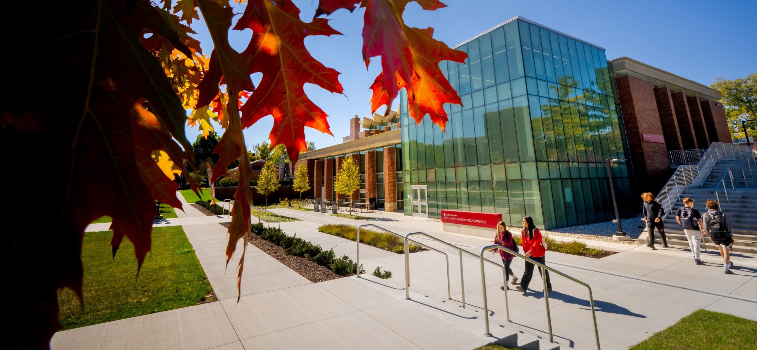 Sunlight shines through autumn leaves beside a modern glass building on the Alma College campus as people walk along a paved path under a clear blue sky, looking forward to enrollment opportunities for Fall 2026.