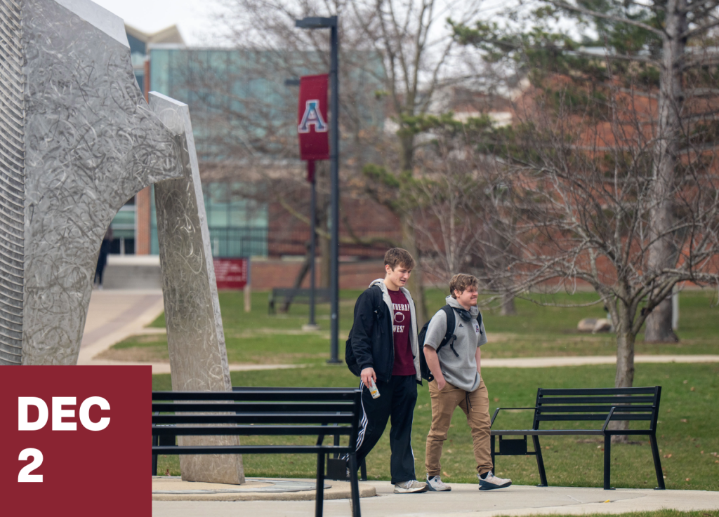 Two students, one a recent transfer, walk on a college campus near benches and a large sculpture. Leafless trees and a building with a red A banner are visible in the background. A box in the corner reads Dec 2.